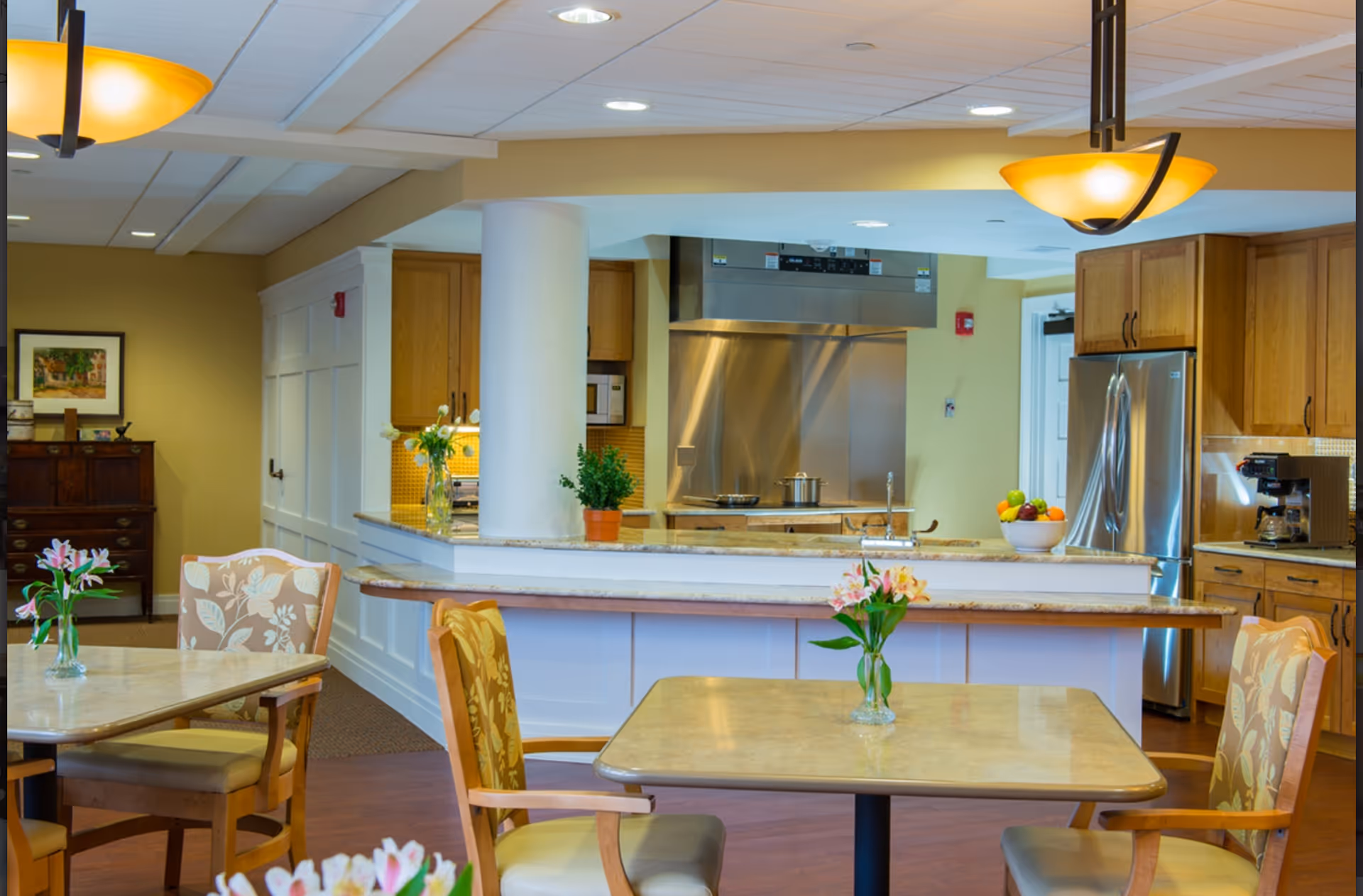 A bright and clean dining area with tables and chairs, each table adorned with a small vase of flowers. In the background, there is a kitchen area with wooden cabinets, a stainless steel refrigerator, a stove with a large stainless steel hood, and a coffee machine. The space has warm lighting and a welcoming atmosphere.