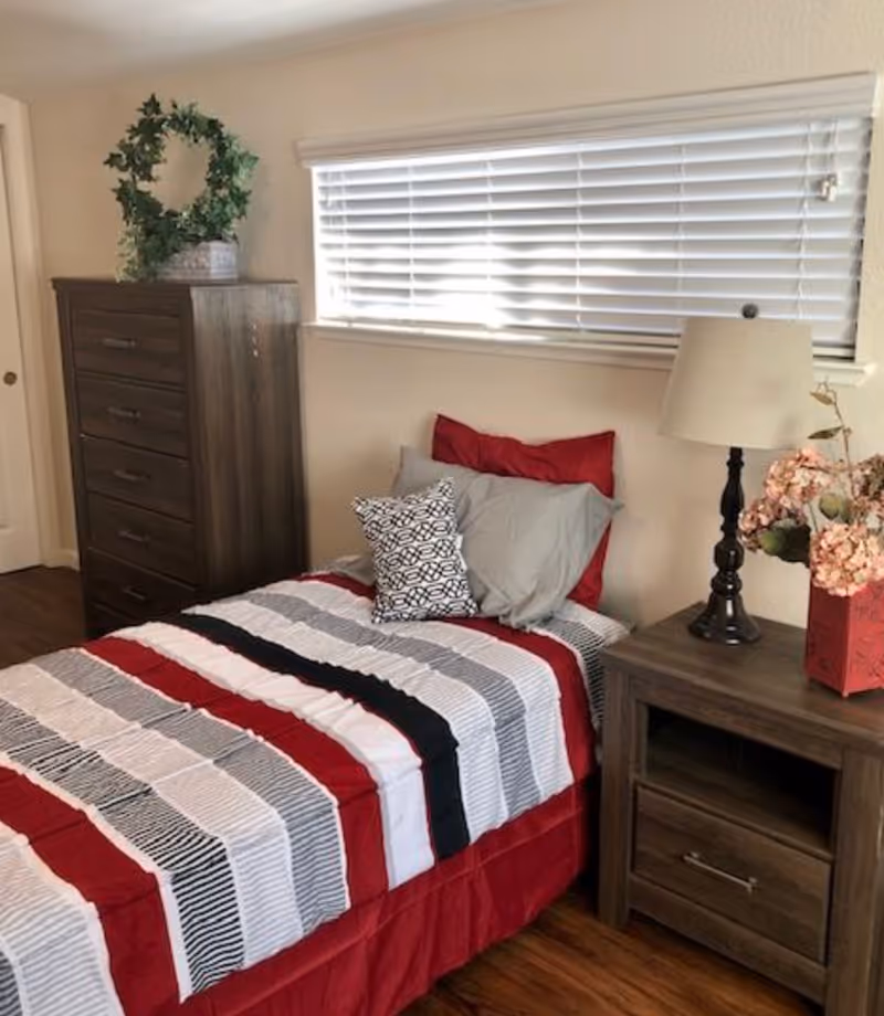 A bedroom with a single bed featuring a striped red, white, black, and gray bedspread and multiple pillows. Next to the bed is a wooden nightstand with a lamp and a vase of pink flowers. A tall wooden dresser with a green leafy wreath on top is against the wall. A window with white blinds is above the bed.