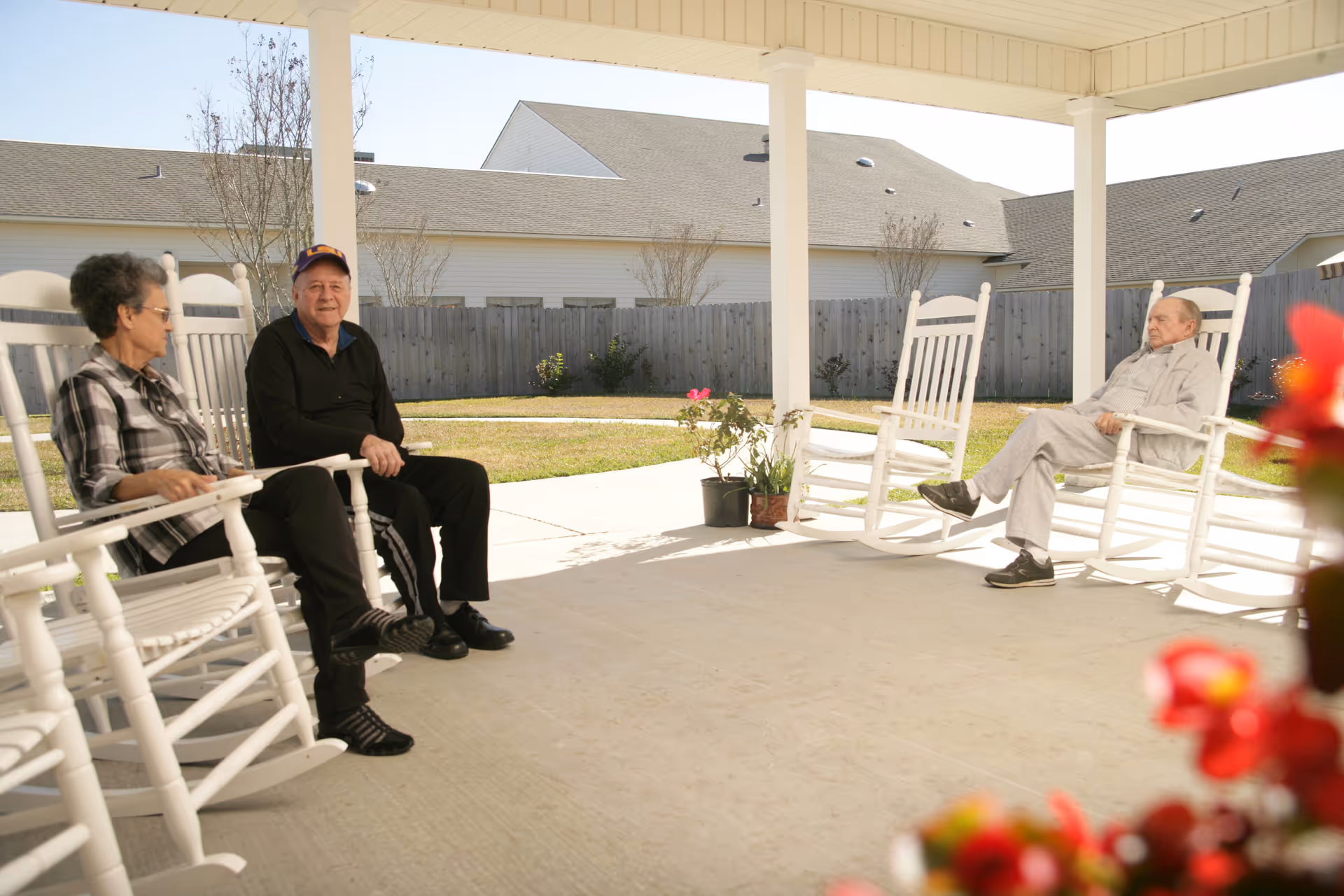 Three elderly individuals sitting on white rocking chairs under a covered patio area with a fenced yard and houses in the background. There are some potted plants on the patio and red flowers in the foreground.