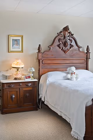 A bedroom with an ornate wooden headboard bed covered with a white bedspread and a white teddy bear sitting on it. Next to the bed is a wooden nightstand with a vintage lamp, a small clock, and other decorative items. A framed portrait hangs on the wall above the nightstand.