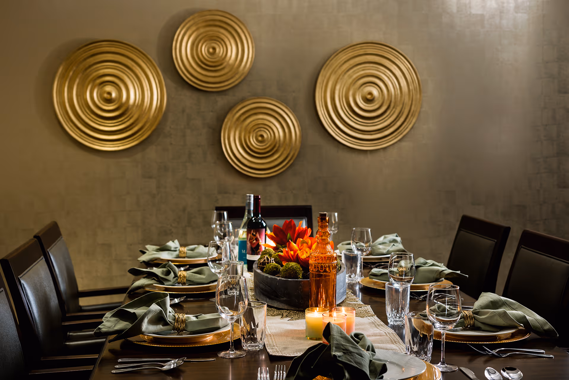 A dining table set for a meal with plates, green cloth napkins with gold napkin rings, wine glasses, water glasses, silverware, and a centerpiece featuring orange flowers and decorative moss balls. There are also two lit candles and bottles of wine and an amber bottle on the table. The background wall has four large decorative gold circular wall hangings.