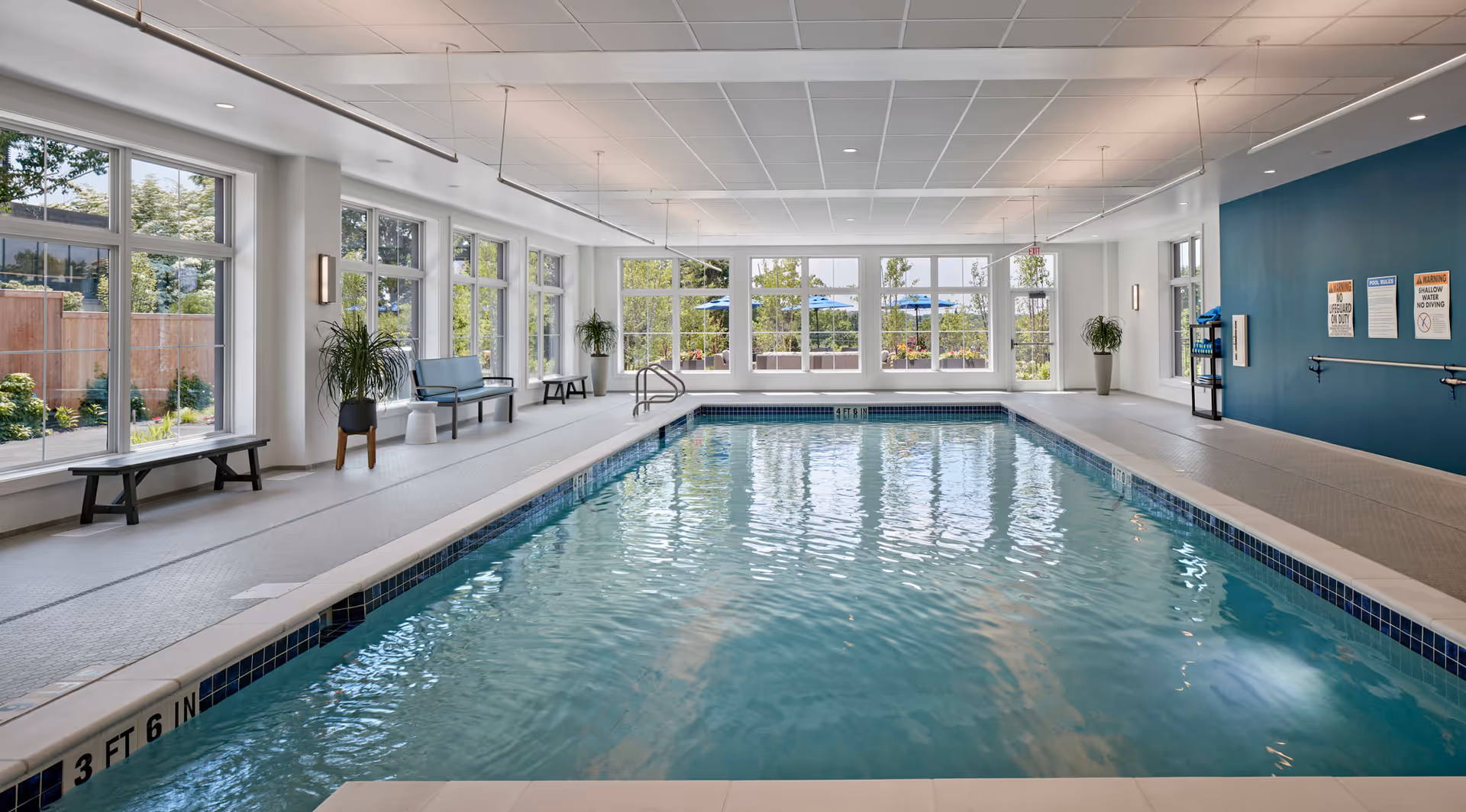 Indoor swimming pool with clear water, surrounded by large windows letting in natural light. There are benches and potted plants along the sides, and safety signs on a blue wall at the far end of the pool area.