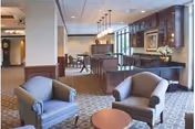 Spacious senior living common area with two armchairs and a small table in the foreground and a dining/bar area with cabinetry in the background.