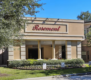 Front exterior view of the Rosemont Assisted Living And Memory Care building with a beige facade, columns at the entrance, greenery, and a clear blue sky.