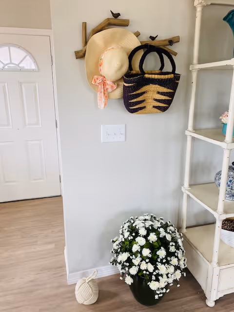 Interior corner of a room with a white door on the left, a wooden wall-mounted coat rack holding a straw hat with a pink ribbon and a woven handbag, a white shelving unit on the right with decorative items, and a large pot of white flowers on the floor.