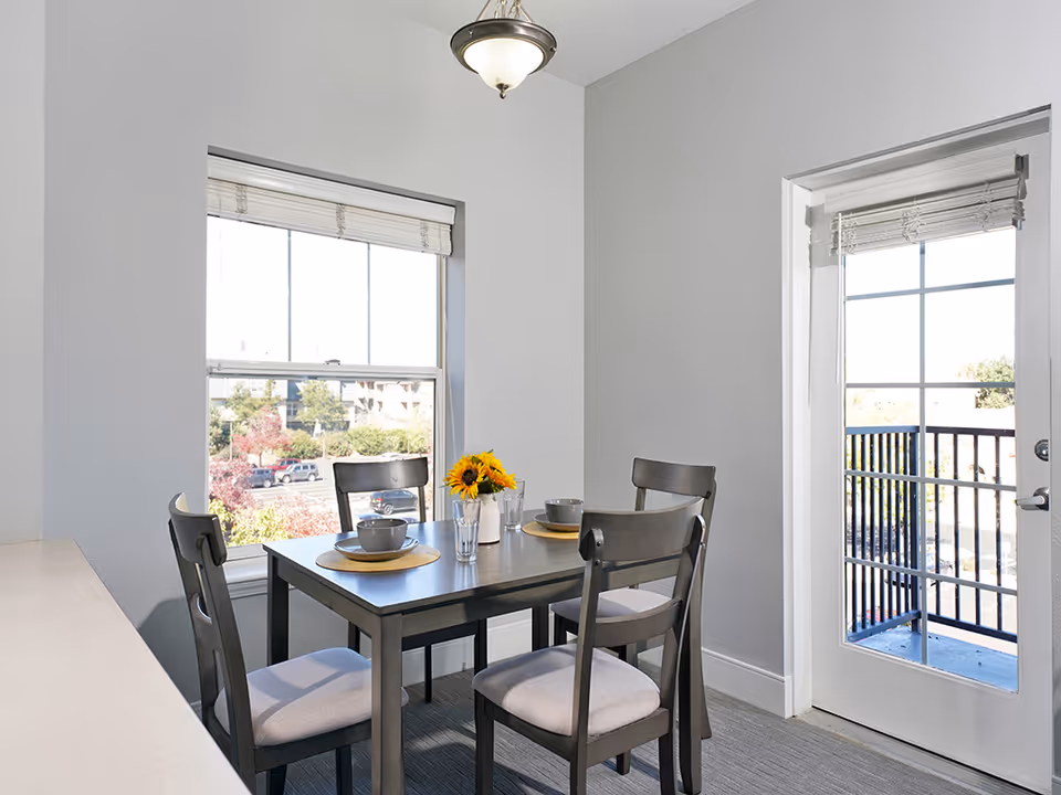 A small dining area with a square dark wooden table set for four people. The table has four matching chairs with light-colored cushions. On the table, there are placemats, bowls, glasses, and a small vase with sunflowers. The room has light gray walls, a window with a view of trees and parked cars outside, and a glass door leading to a balcony with a black railing.