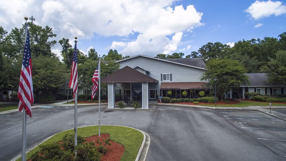 Front exterior view of Oaks at Savannah facility with a driveway, three American flags on flagpoles, landscaped greenery, and a partly cloudy sky.