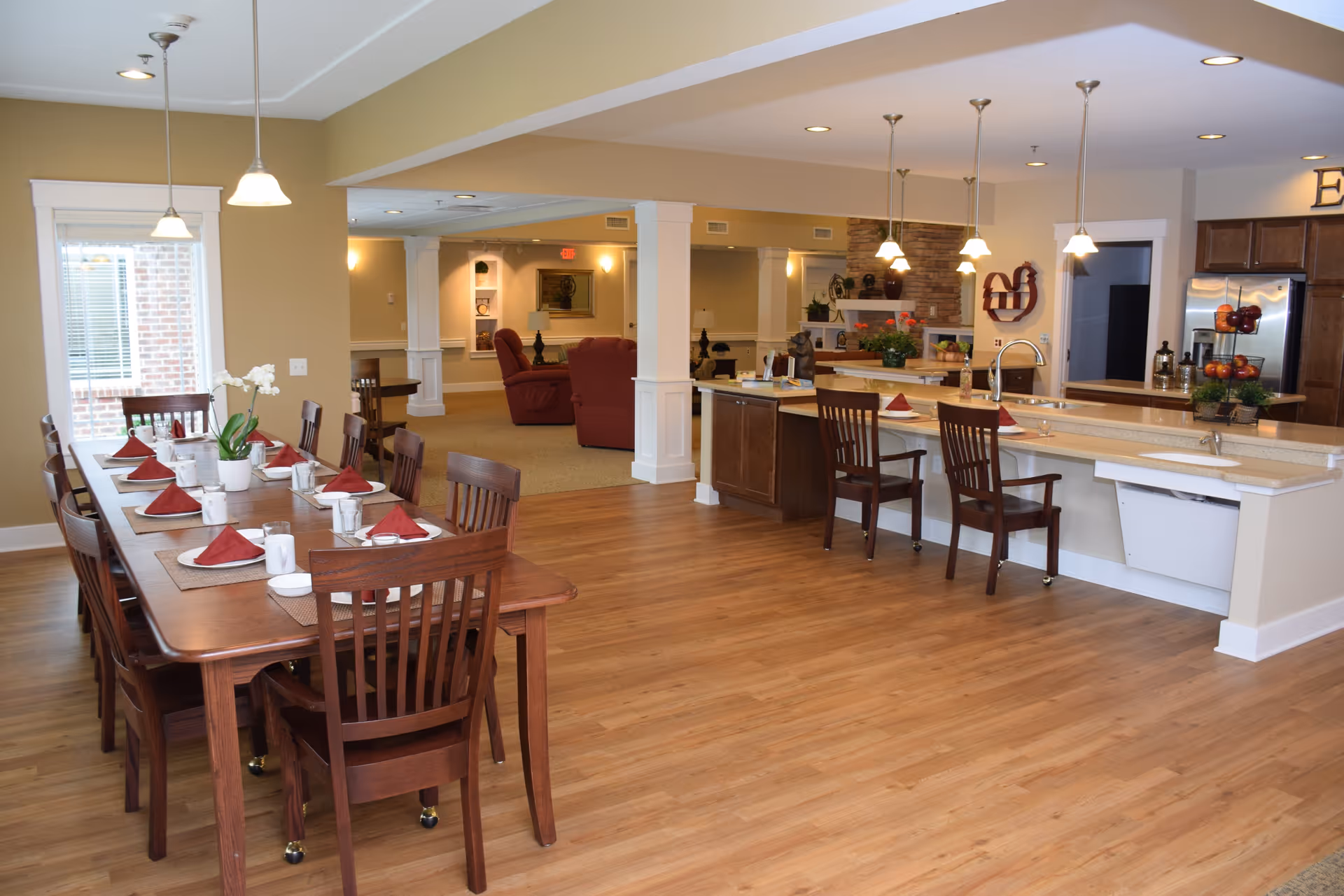 A spacious senior living facility interior featuring a dining area with a long wooden table set with red napkins, white cups, and placemats. Adjacent to the dining area is a kitchen with a large island, bar stools, pendant lighting, and stainless steel appliances. In the background, a living room area with red upholstered chairs and a fireplace is visible.