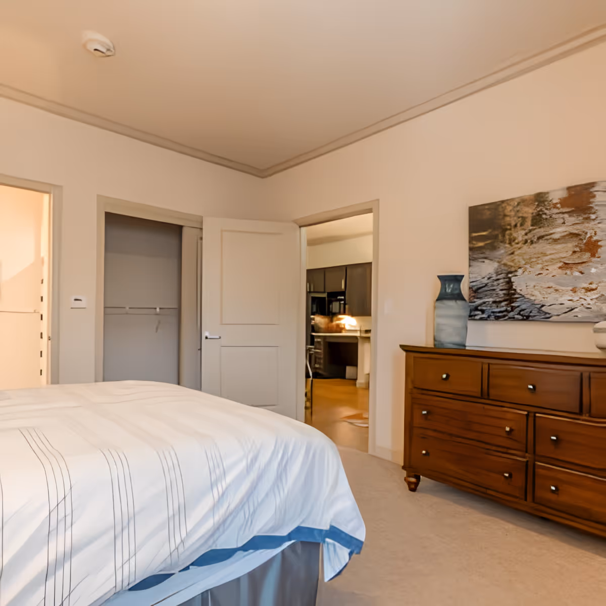A bedroom with a neatly made bed covered in a white and blue striped bedspread. There is a wooden dresser with multiple drawers on the right side, topped with a blue vase and a piece of abstract artwork hanging above it. Two open doors are visible, one leading to a closet and the other to a kitchen area with dark cabinets and a countertop.