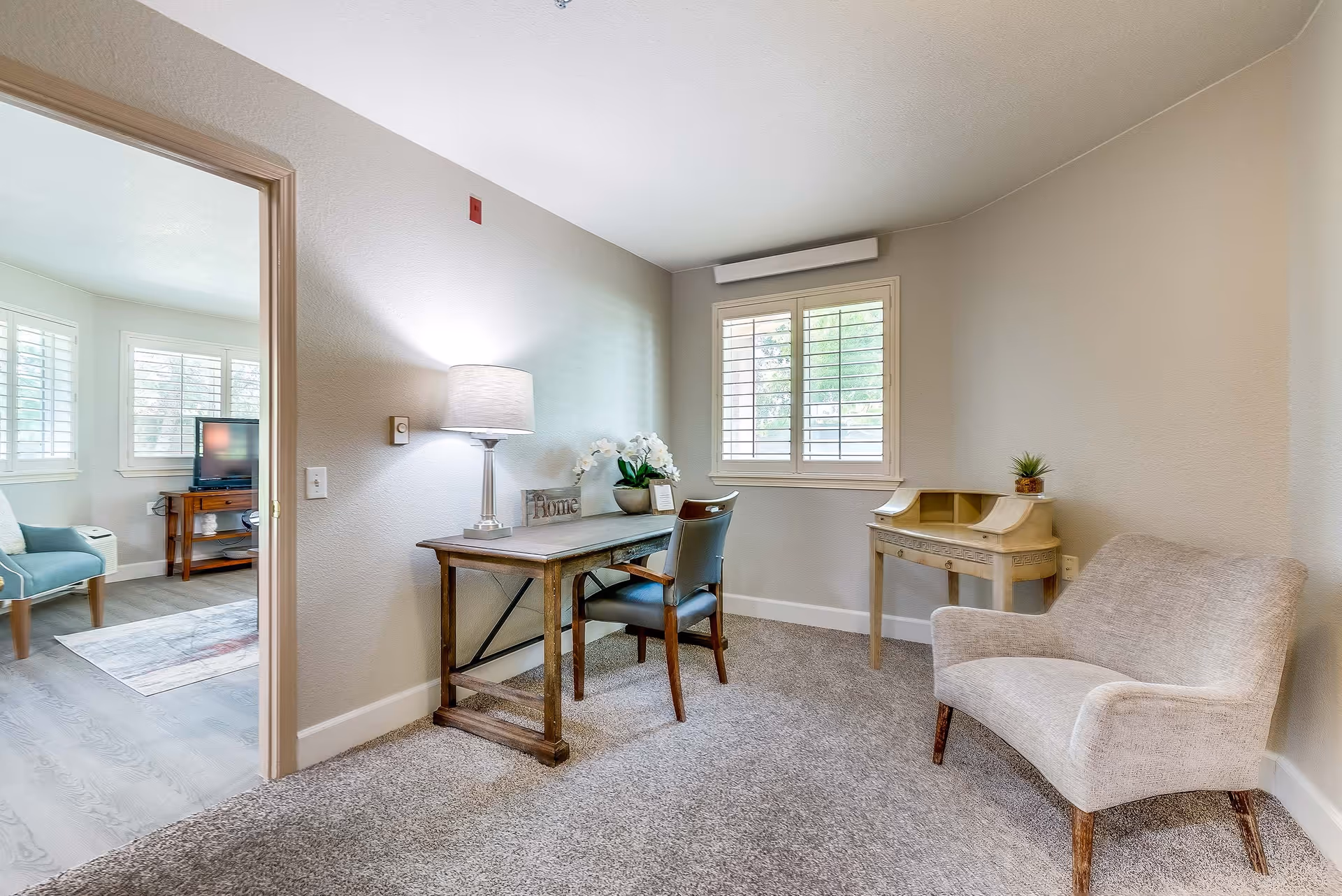 A cozy interior room with a wooden desk and chair, an upholstered armchair, and a window with shutters.