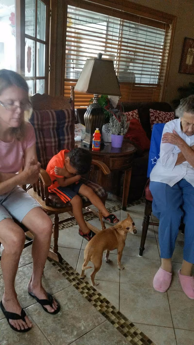 An elderly woman, a young boy, and a middle-aged woman sitting in a living room with a small brown dog standing on the tiled floor. The elderly woman is wearing blue pants and pink slippers, the boy is dressed in an orange shirt and shorts, and the middle-aged woman is wearing a pink shirt and shorts. There is a wooden side table with a lamp, a purple flower pot, and a bottle on it, with a window with blinds in the background.