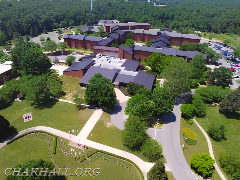 Aerial view of the Charlotte Hall Veterans Home facility surrounded by green trees and lawns, showing multiple connected buildings with dark roofs and pathways leading to the entrance.