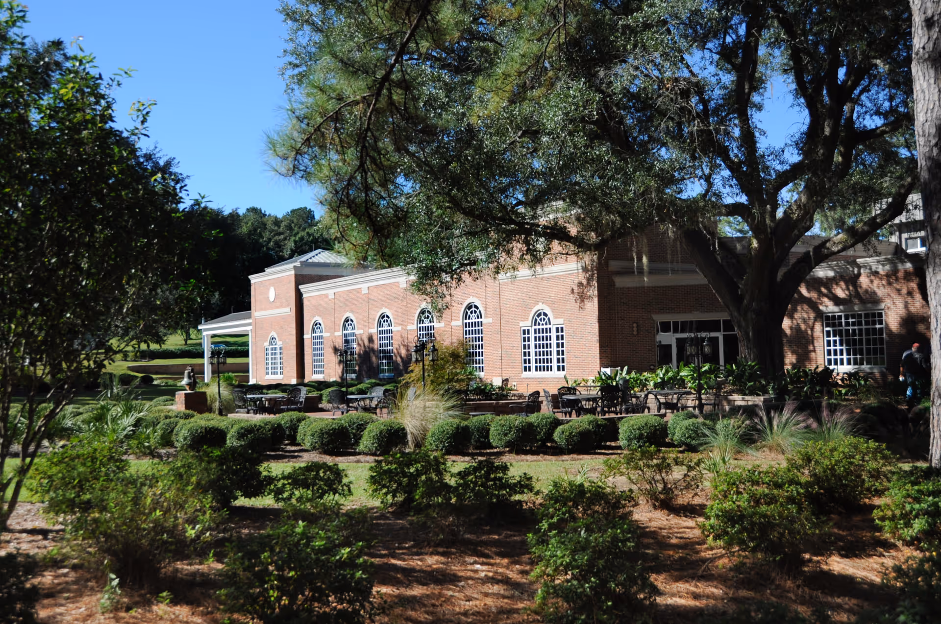 A brick building with large arched windows surrounded by a landscaped garden with bushes, trees, and outdoor seating under a clear blue sky.