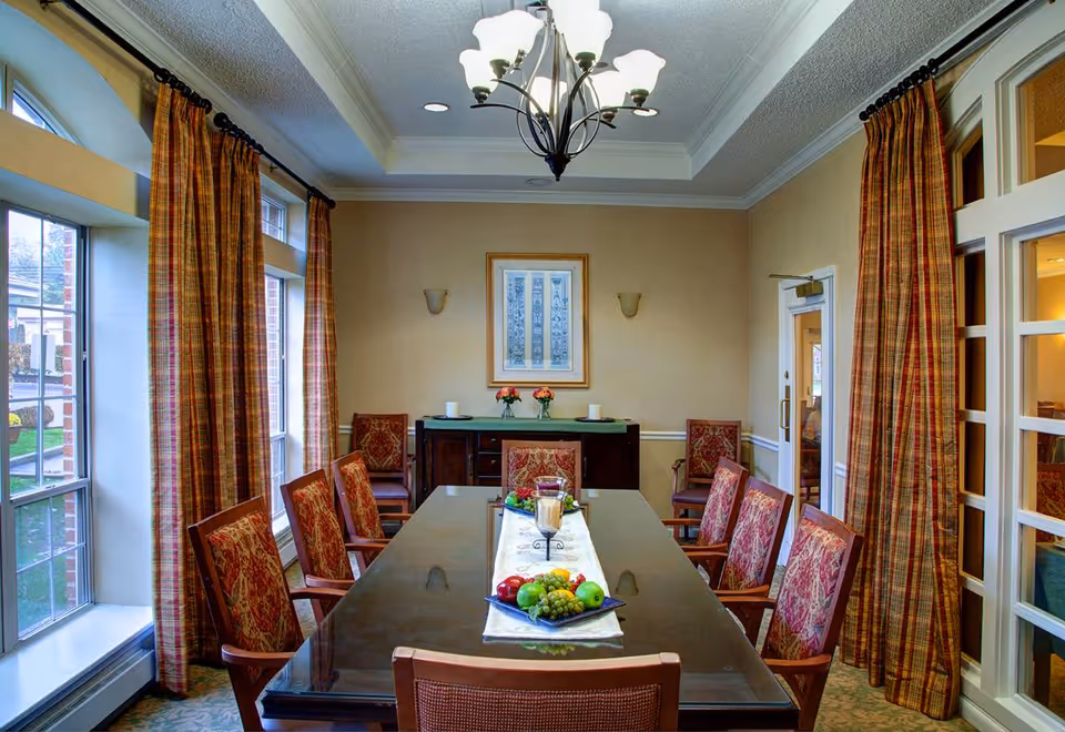 A formal dining room with a long polished table surrounded by upholstered chairs, plaid curtains, a chandelier, and large windows.