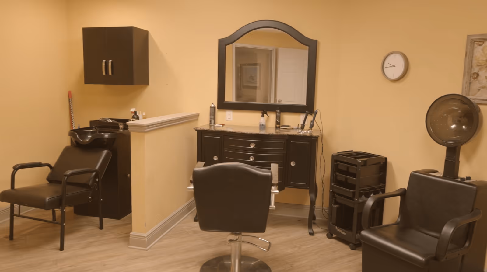 Interior of a hair salon area with a styling chair in front of a vanity with a mirror, a black salon sink with a chair, a hair dryer chair, and a rolling cart with hair styling tools. The walls are painted beige and there is a clock on the wall.