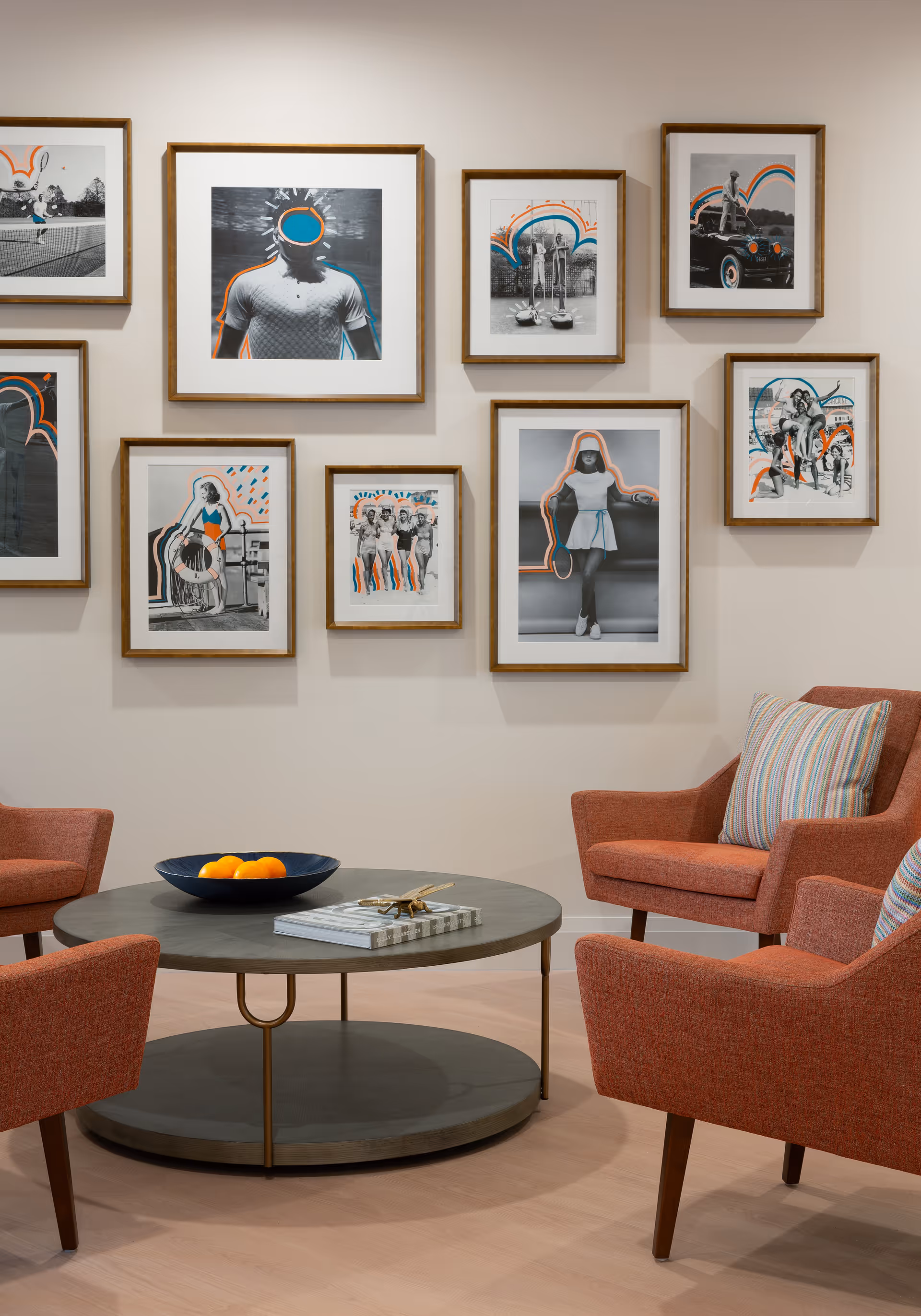 A cozy seating area with four orange upholstered armchairs arranged around a round coffee table. The table has a bowl of oranges and a stack of books on it. The wall behind the chairs features a gallery of framed black-and-white photographs with colorful abstract outlines and accents.