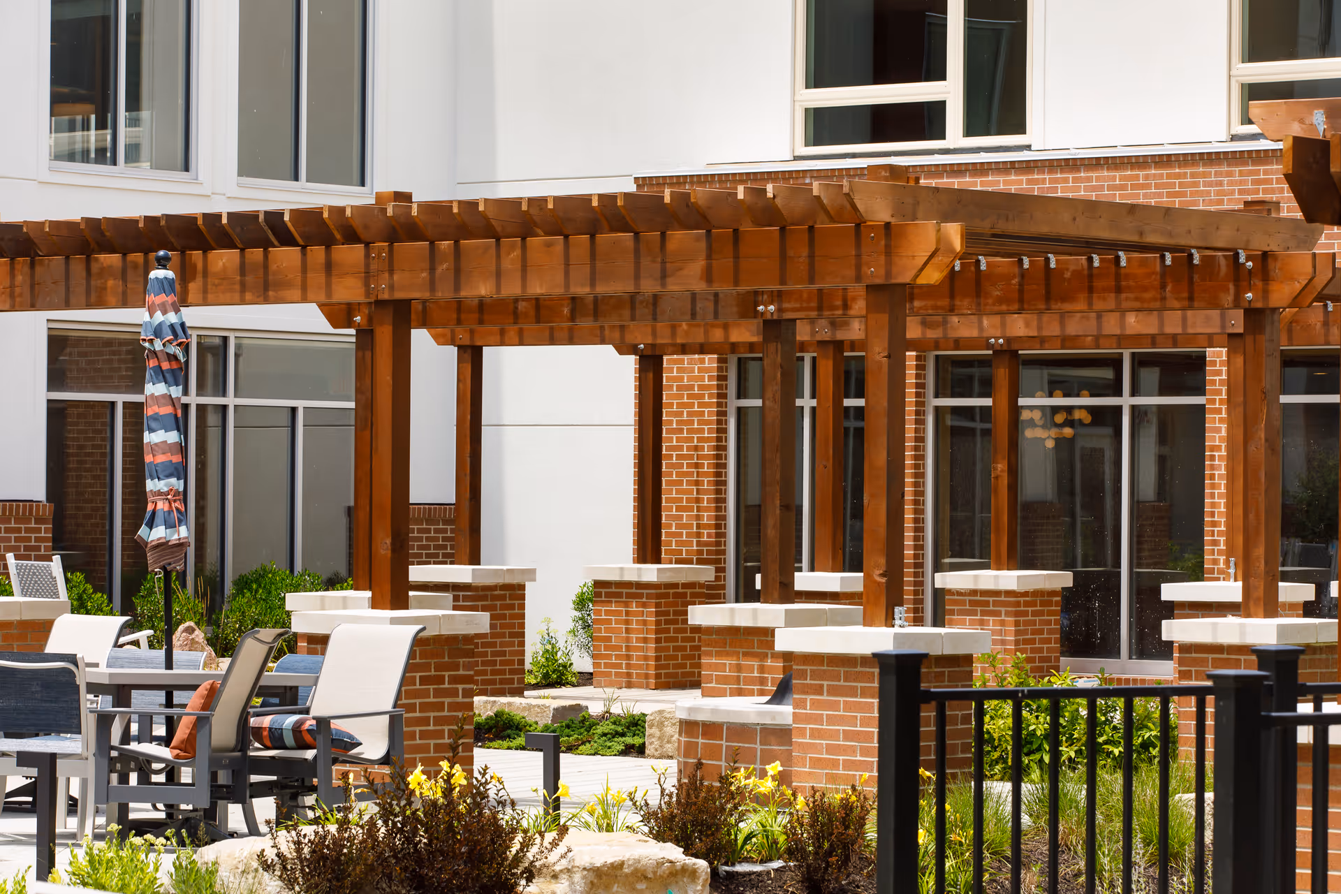 Outdoor patio area with wooden pergolas, brick pillars, tables, chairs, and a closed striped umbrella, surrounded by plants and greenery in front of a building with large windows.