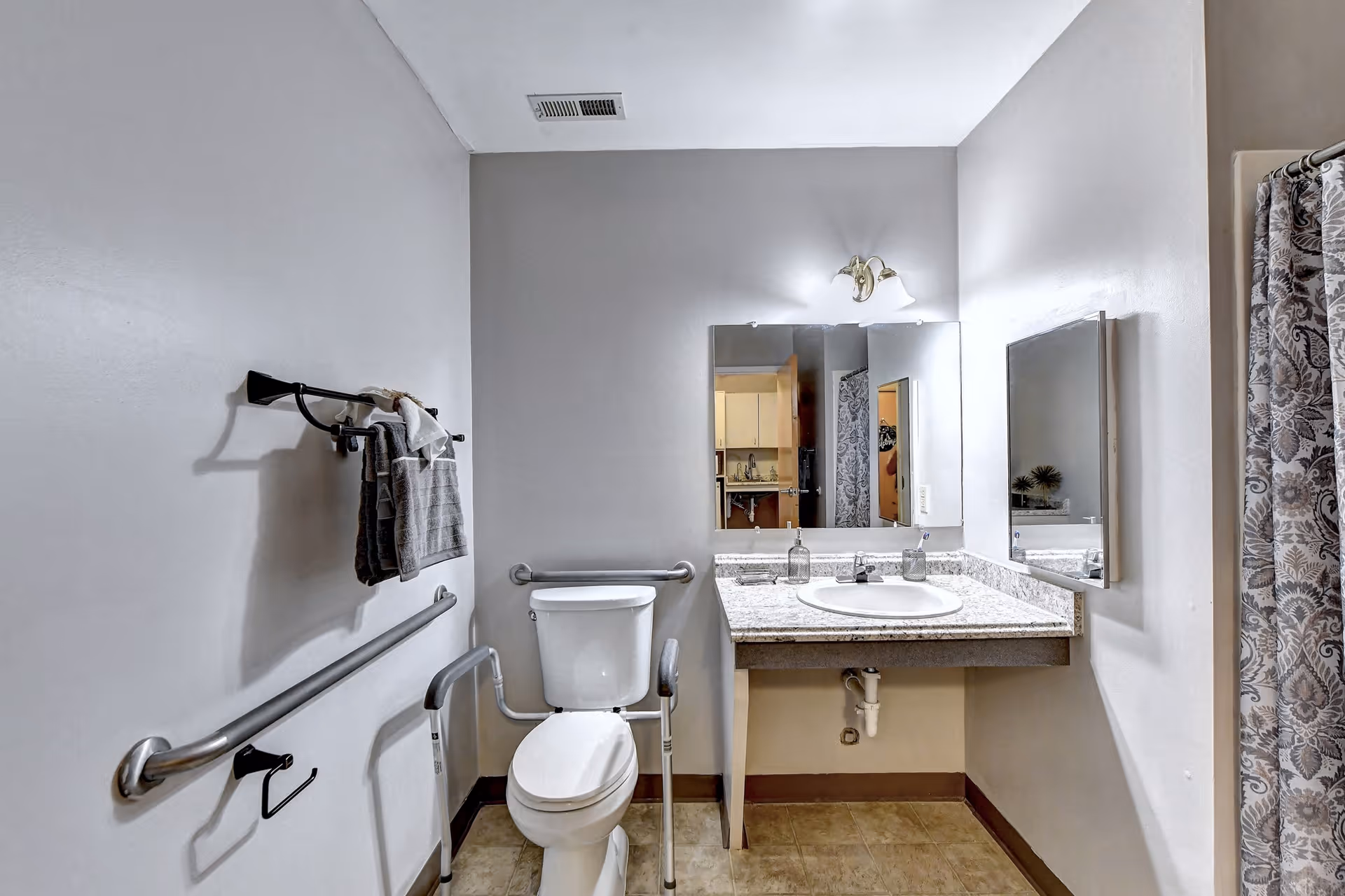 Accessible bathroom with a toilet, grab bars, sink and mirror, towel rack, and a patterned shower curtain.