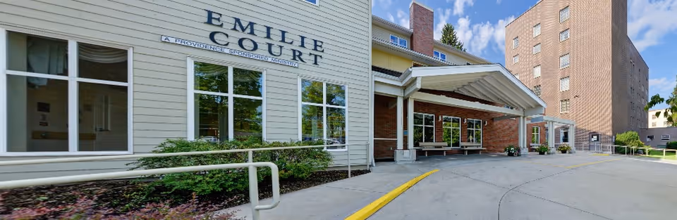 Exterior view of Emilie Court senior living facility showing the building entrance with a covered drop-off area, windows, and a ramp with handrails. The sky is partly cloudy and there are some plants along the building.