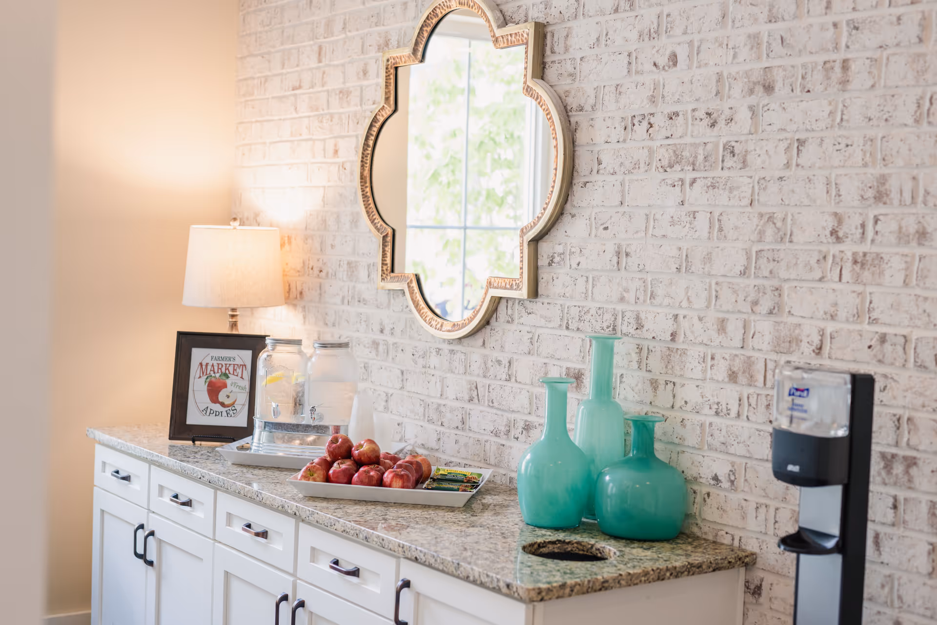 A countertop with white cabinets below, decorated with a lamp, a framed sign that says 'Farmer's Market Fresh Apples', two glass beverage dispensers with water and lemon slices, a tray of red apples, three turquoise decorative vases, and a wall-mounted mirror with a decorative frame. There is also a hand sanitizer dispenser on the right side against a white brick wall.