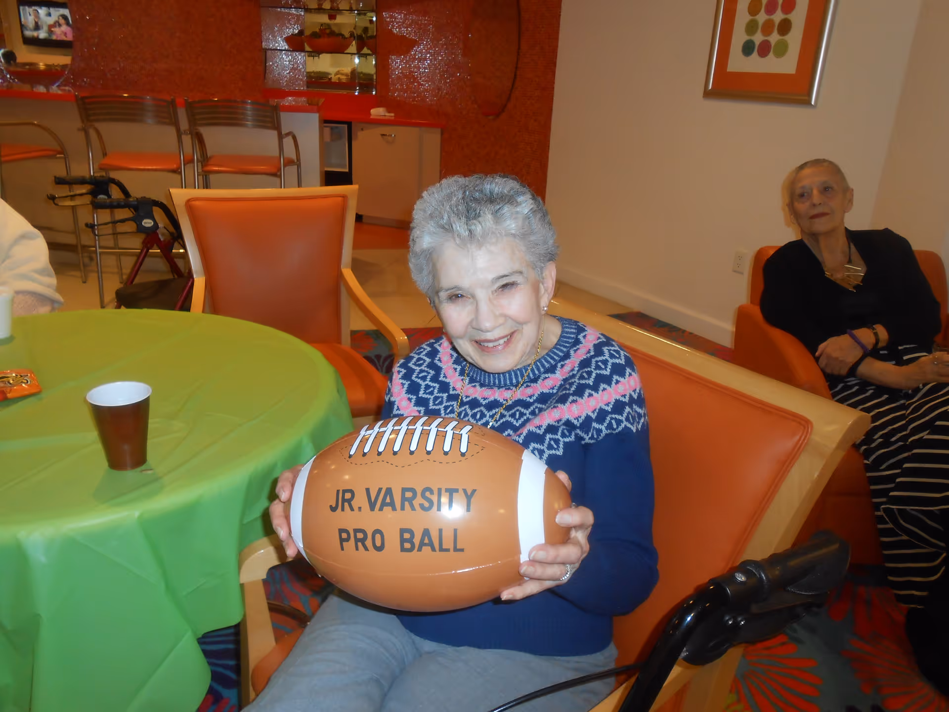An elderly woman sitting in an orange chair indoors, smiling and holding a brown football labeled 'JR. VARSITY PRO BALL'. Another elderly woman is seated in the background on an orange chair. The room has a green tablecloth on a round table and orange chairs, with a kitchen area visible in the background.