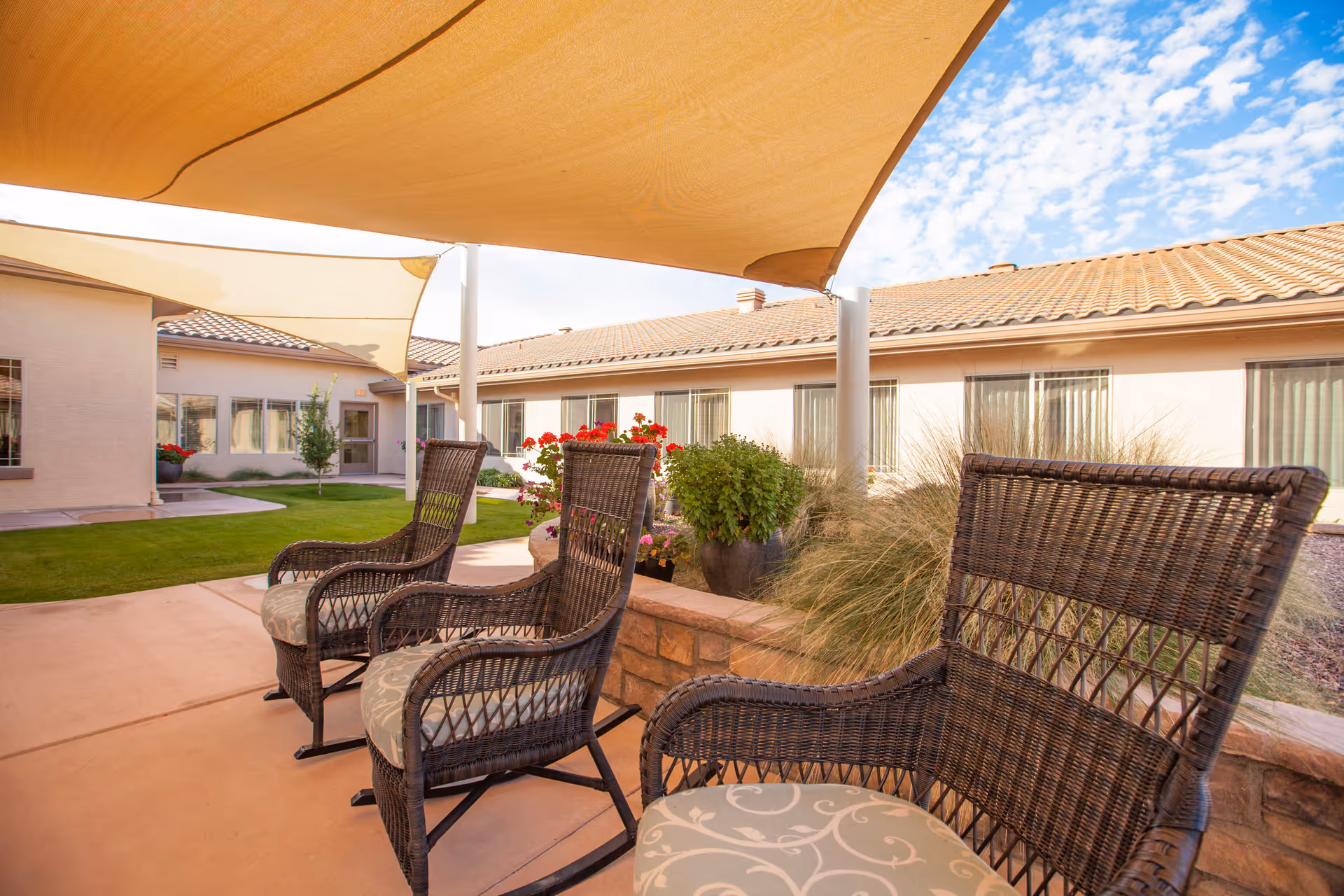 Outdoor seating area with three wicker chairs with cushions under large beige shade sails. The area is paved and surrounded by a low stone wall with potted plants and flowers. In the background, there is a single-story building with multiple windows and a tiled roof under a partly cloudy sky.