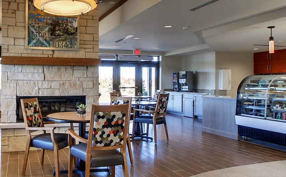 A cozy dining area in Evergreen Senior Living featuring round wooden tables with patterned cushioned chairs, a stone fireplace with artwork above it, a beverage dispenser, and a glass display case with snacks. The space has wooden flooring and large windows letting in natural light.