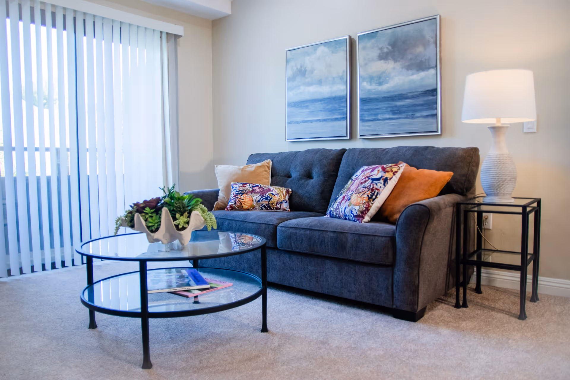 Living room with a gray upholstered sofa topped with colorful throw pillows, a round glass coffee table with a plant, framed seascape art, and a lamp by sliding vertical blinds.