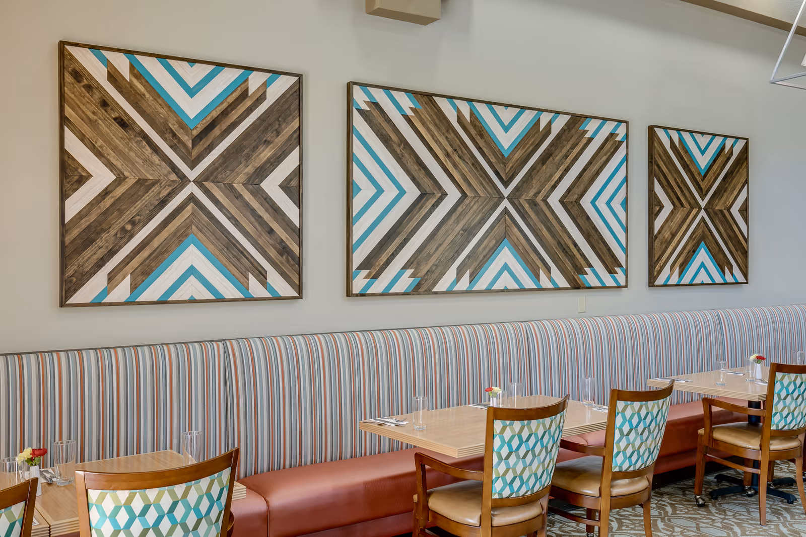 A dining area with a long striped upholstered bench along the wall, wooden tables set with glasses and small flower vases, and wooden chairs with patterned backs. Three large geometric wooden art pieces with blue, white, and brown colors hang on the wall above the bench.