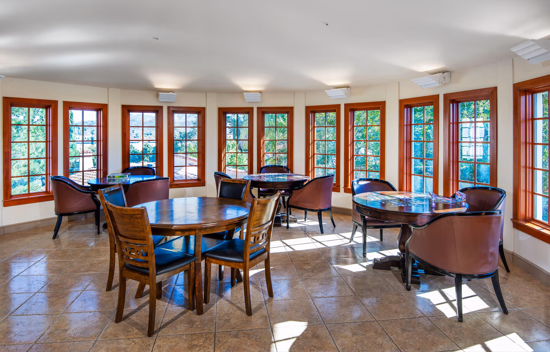 Bright communal dining/activity room with round wooden tables and leather chairs surrounded by tall multi-pane windows.