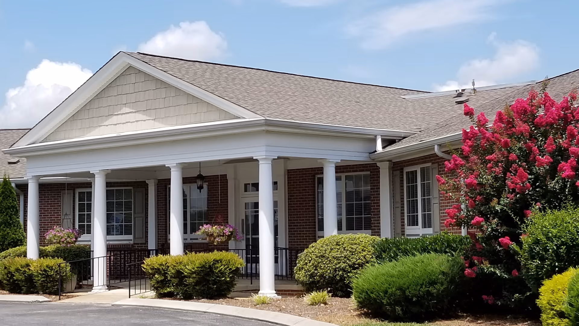 Front entrance of a brick building with a covered porch supported by white columns, trimmed hedges, and a blooming pink flowering shrub.