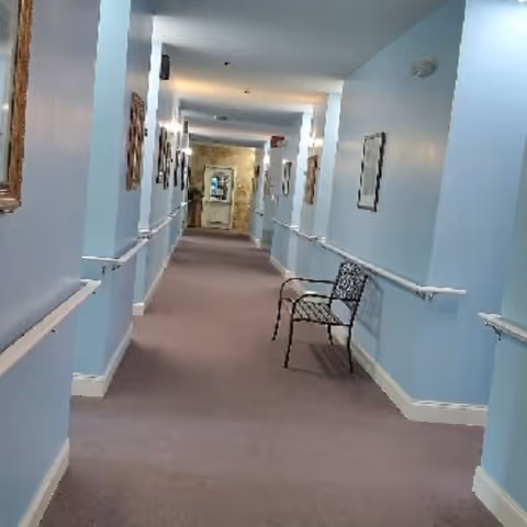A long light-blue hallway in an assisted living facility with handrails, framed artwork, and a metal bench.