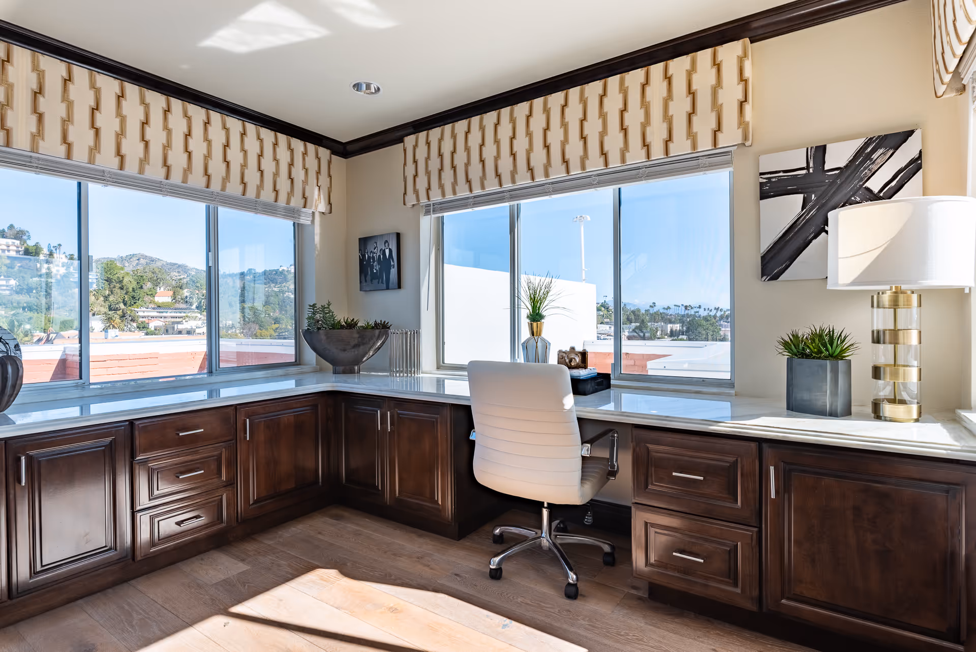 A bright corner office space with large windows offering a view of hills and buildings outside. The room features dark wooden cabinets with a white marble countertop, a white swivel office chair, decorative plants, a modern table lamp, and abstract wall art.