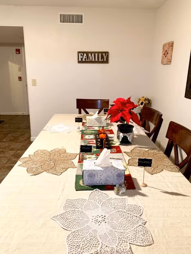 A dining table set for a meal with decorative placemats shaped like flowers, a red poinsettia plant centerpiece, tissue boxes, and small name cards labeled John and Michael. The wall behind the table has a sign that says FAMILY and a small decorative piece. The room has wooden chairs and tiled flooring.