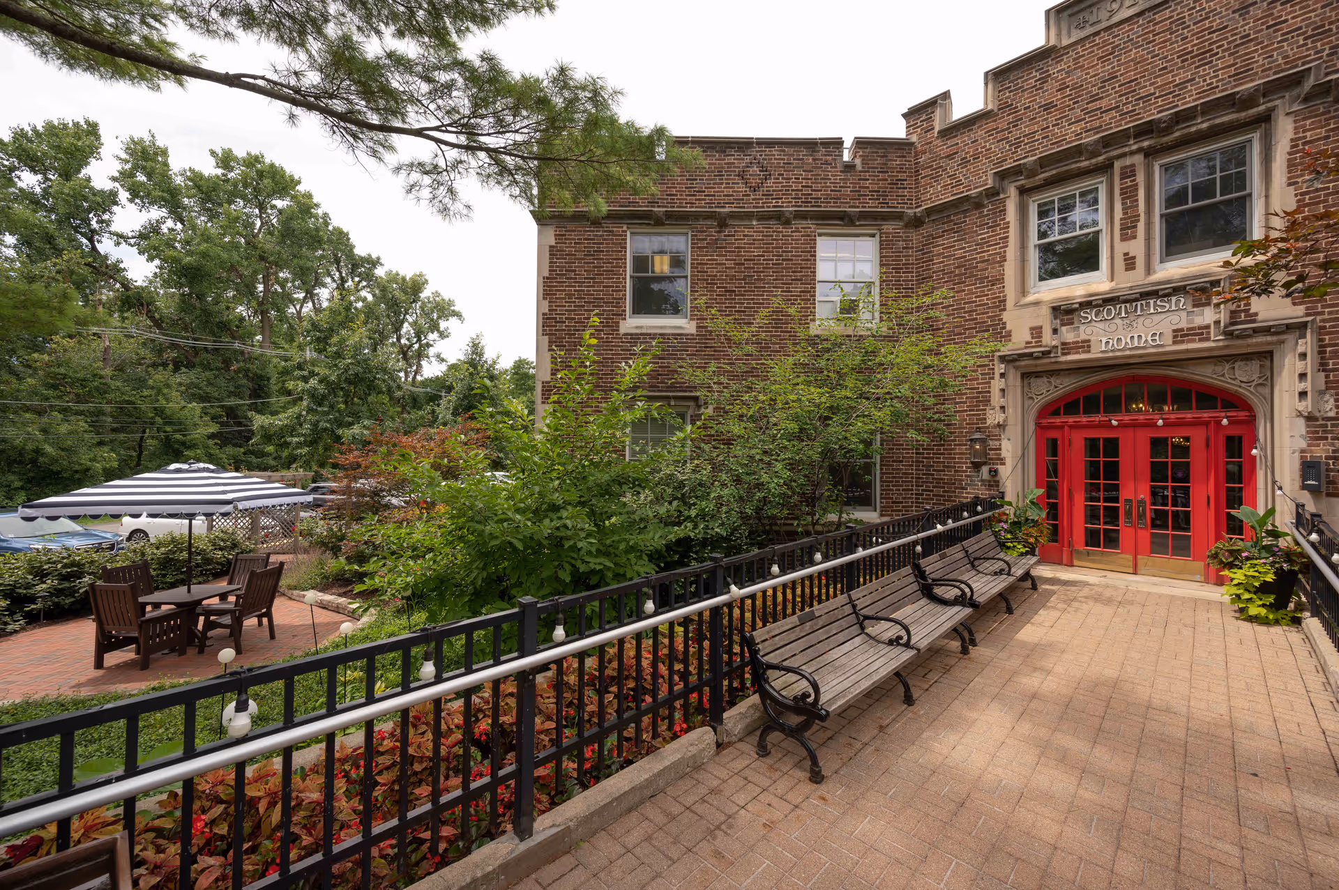 Outdoor view of a senior living facility entrance with a brick pathway, black metal railing, wooden benches, and a garden area with green shrubs and plants. The building has a brick facade with red double doors under an archway labeled 'Scottish Home'. There is a patio area with a table, chairs, and a striped umbrella to the left.
