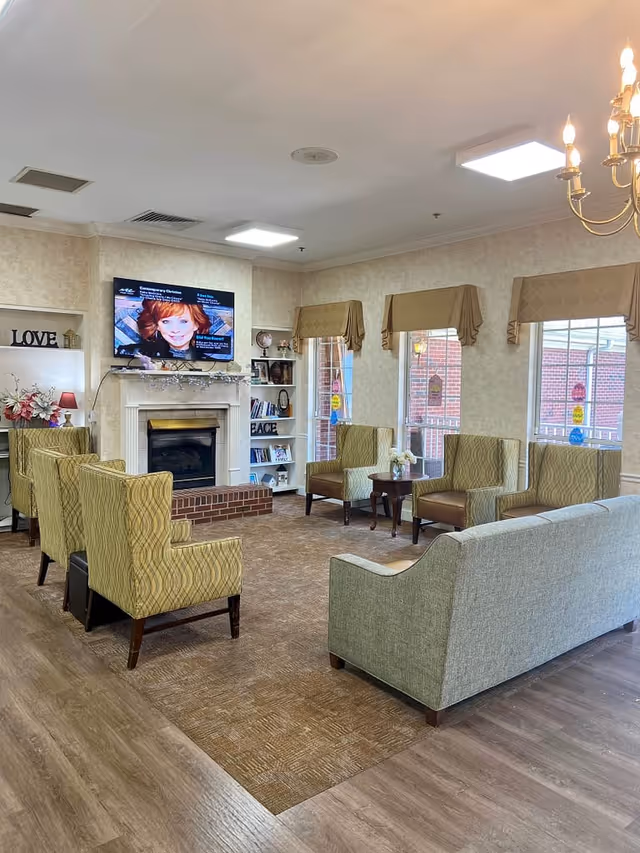 A cozy living room area in a senior living facility with multiple upholstered armchairs and a sofa arranged around a fireplace. Above the fireplace is a mounted flat-screen TV displaying a woman's face. The room has large windows with beige valances, built-in shelves with decorative items and books, and a chandelier hanging from the ceiling. The floor is a combination of wood and carpet.