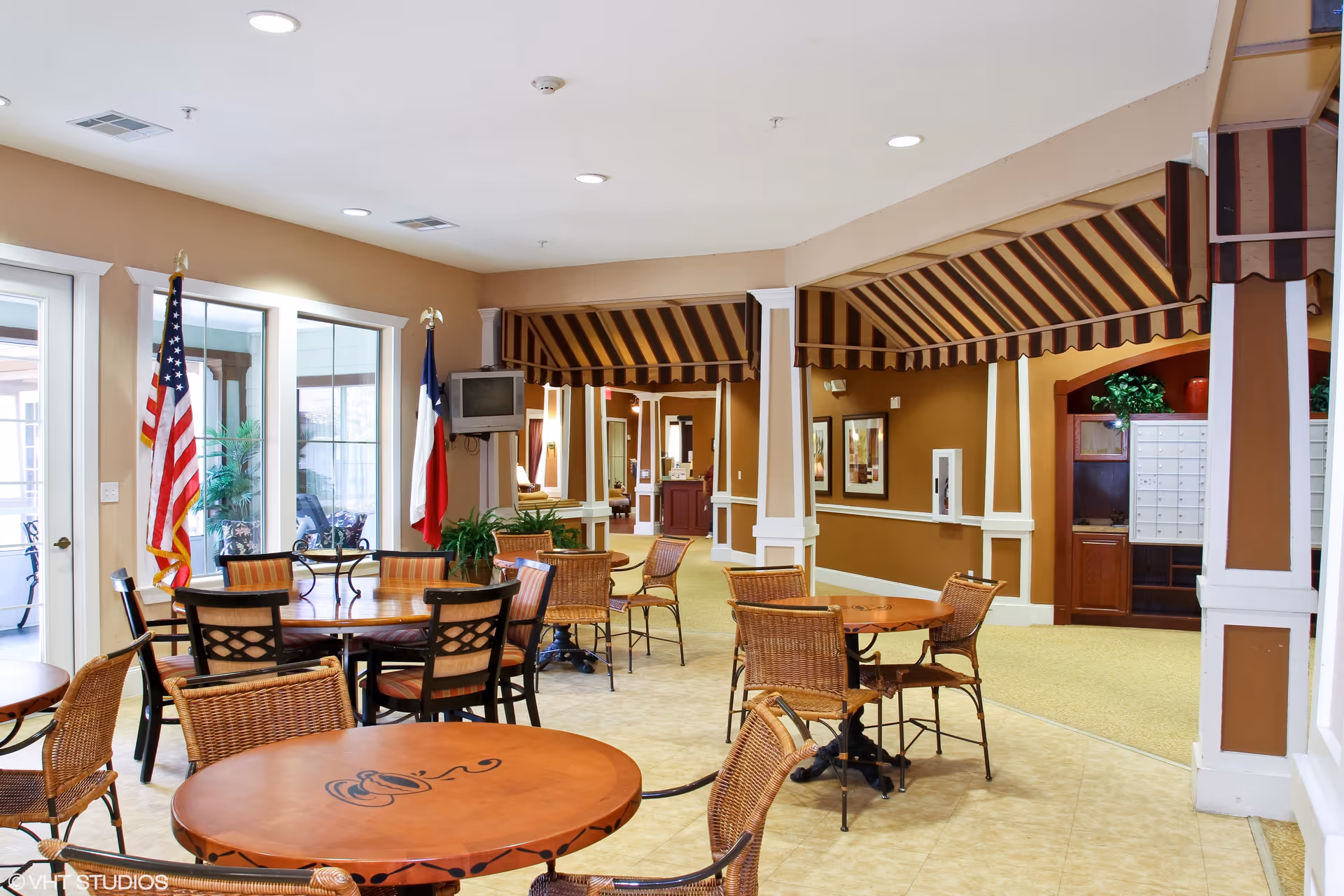 Interior view of a common area in Magnolia Court Assisted Living and Memory Care featuring round wooden tables with wicker and cushioned chairs. The walls are painted brown with white trim, and there are striped awnings above the seating areas. Two flags, an American flag and a Texas state flag, stand near large windows letting in natural light. A small wall-mounted television is visible, along with some plants and framed artwork on the walls.