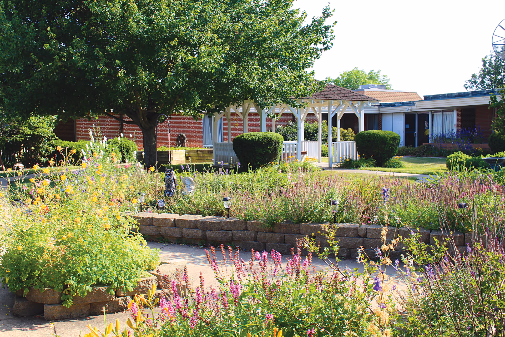 A sunny outdoor garden area at Sunny Acres Nursing Home featuring colorful flowers, green shrubs, a large tree, a white gazebo, and a brick building in the background.