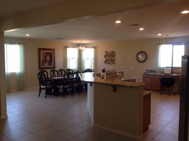 Interior view of a spacious room in an assisted living facility featuring a dining area with a dark wooden table and chairs, a kitchen counter with a granite countertop, and a small office area with a desk and chair near a window. The room has tiled floors, light-colored walls, recessed ceiling lights, and windows with light green curtains allowing natural light to enter.