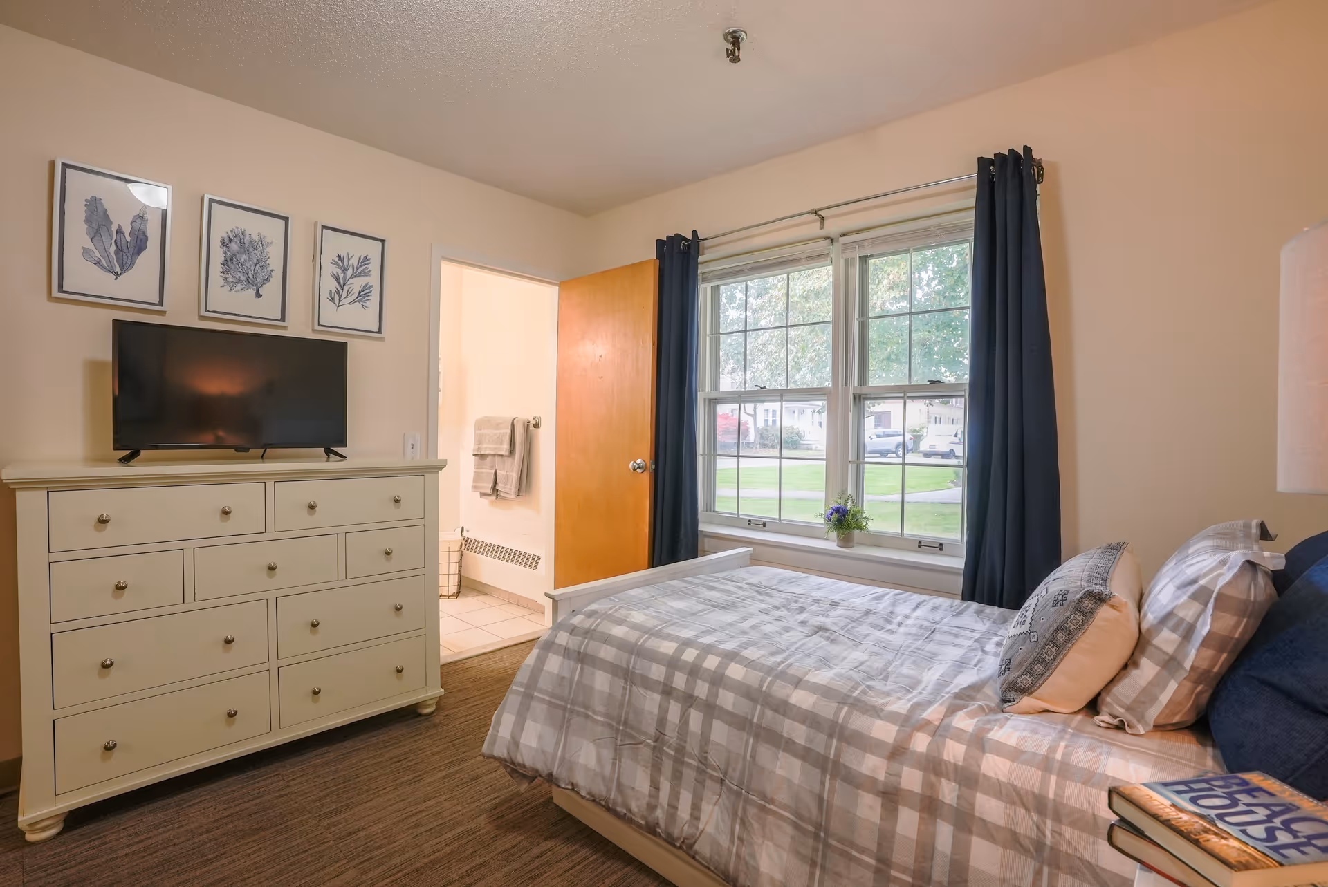 A cozy bedroom with a single bed covered in gray and white checkered bedding and multiple pillows. There is a white dresser with a flat-screen TV on top, and three framed botanical prints hanging above it. A window with dark curtains lets in natural light, showing a green lawn and houses outside. An open door reveals a bathroom with towels hanging on a rack.