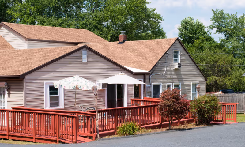 Exterior view of a single-story residential building with beige siding and a brown shingled roof. The building features a red wooden deck with railings, two patio umbrellas, outdoor chairs, and some small shrubs and plants along the deck. Trees and a wooden fence are visible in the background under a partly cloudy sky.