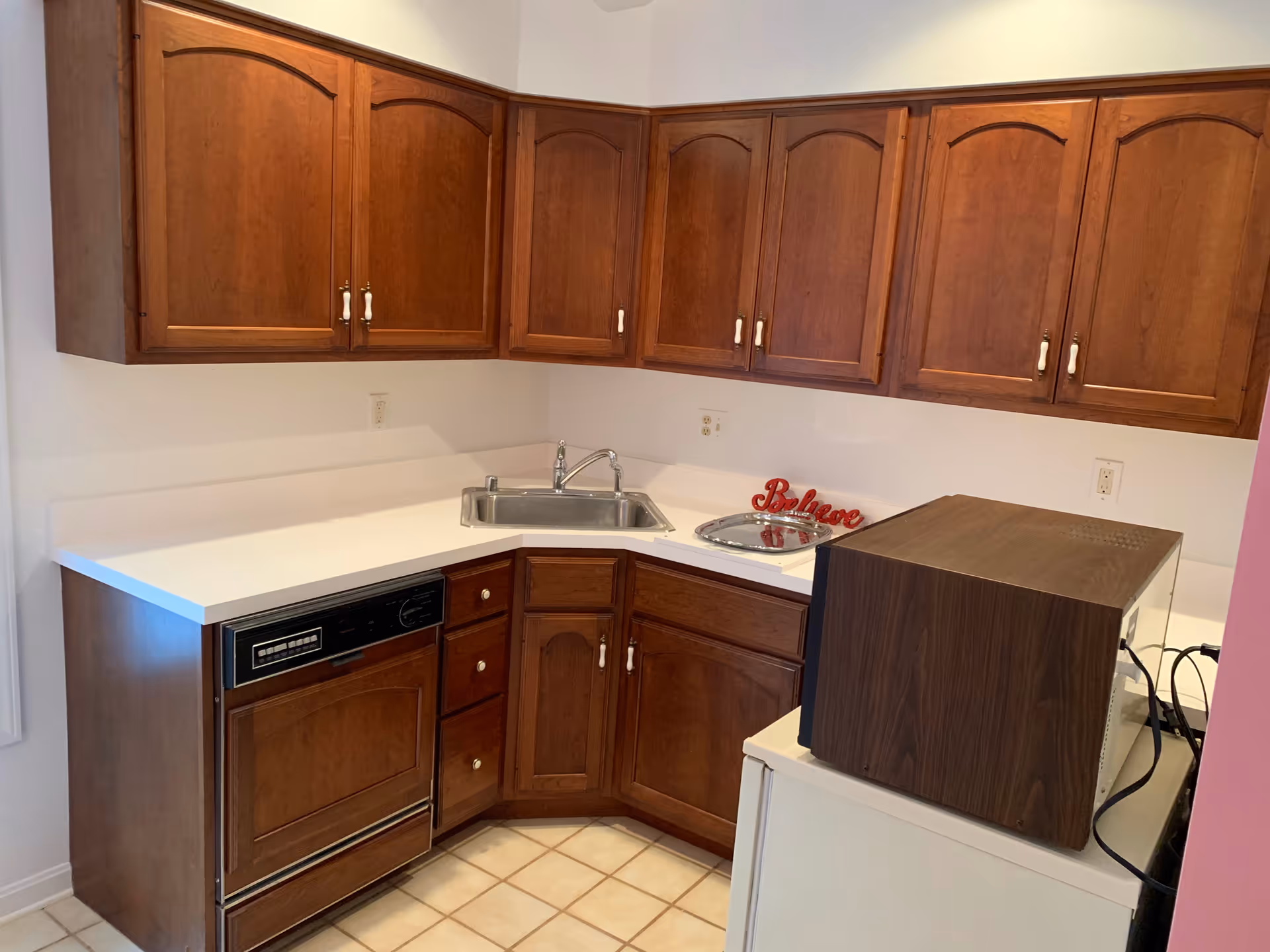 A small kitchen area with wooden cabinets, a white countertop, a stainless steel sink, a built-in dishwasher, and a microwave placed on top of a white appliance. There is a decorative red sign that says 'Believe' on the countertop near the sink.