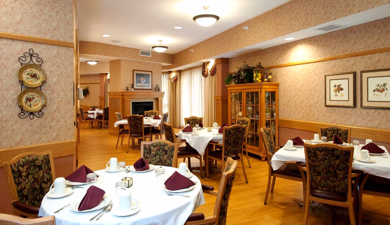 A cozy dining room with several round tables covered in white tablecloths, each set with plates, cups, silverware, and folded burgundy napkins. The room features wooden chairs with patterned upholstery, wood flooring, wallpapered walls, a fireplace, and a glass cabinet with decorative items. Soft lighting and large windows with curtains create a warm atmosphere.