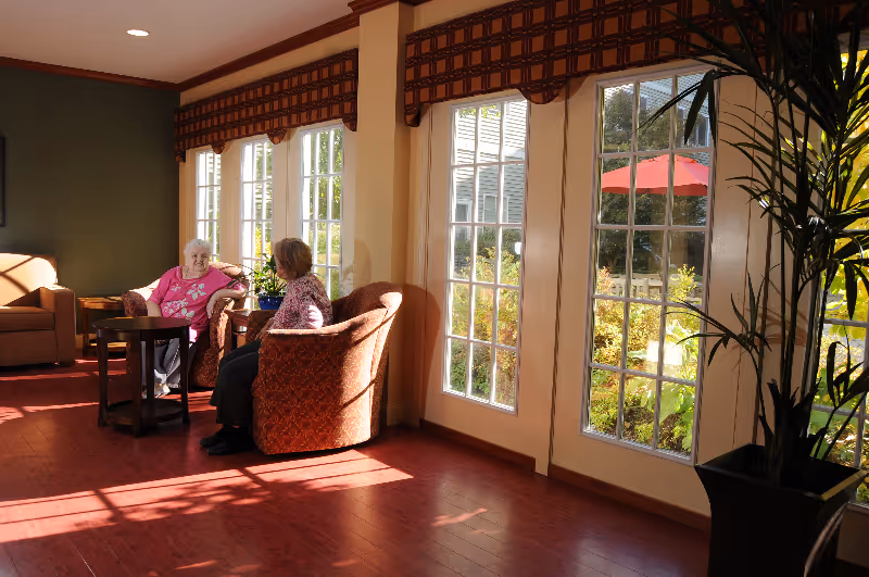 Two elderly women sitting and talking in a sunlit common area with large windows showing a garden outside. The room has wooden floors, comfortable armchairs, and a potted plant near the windows.