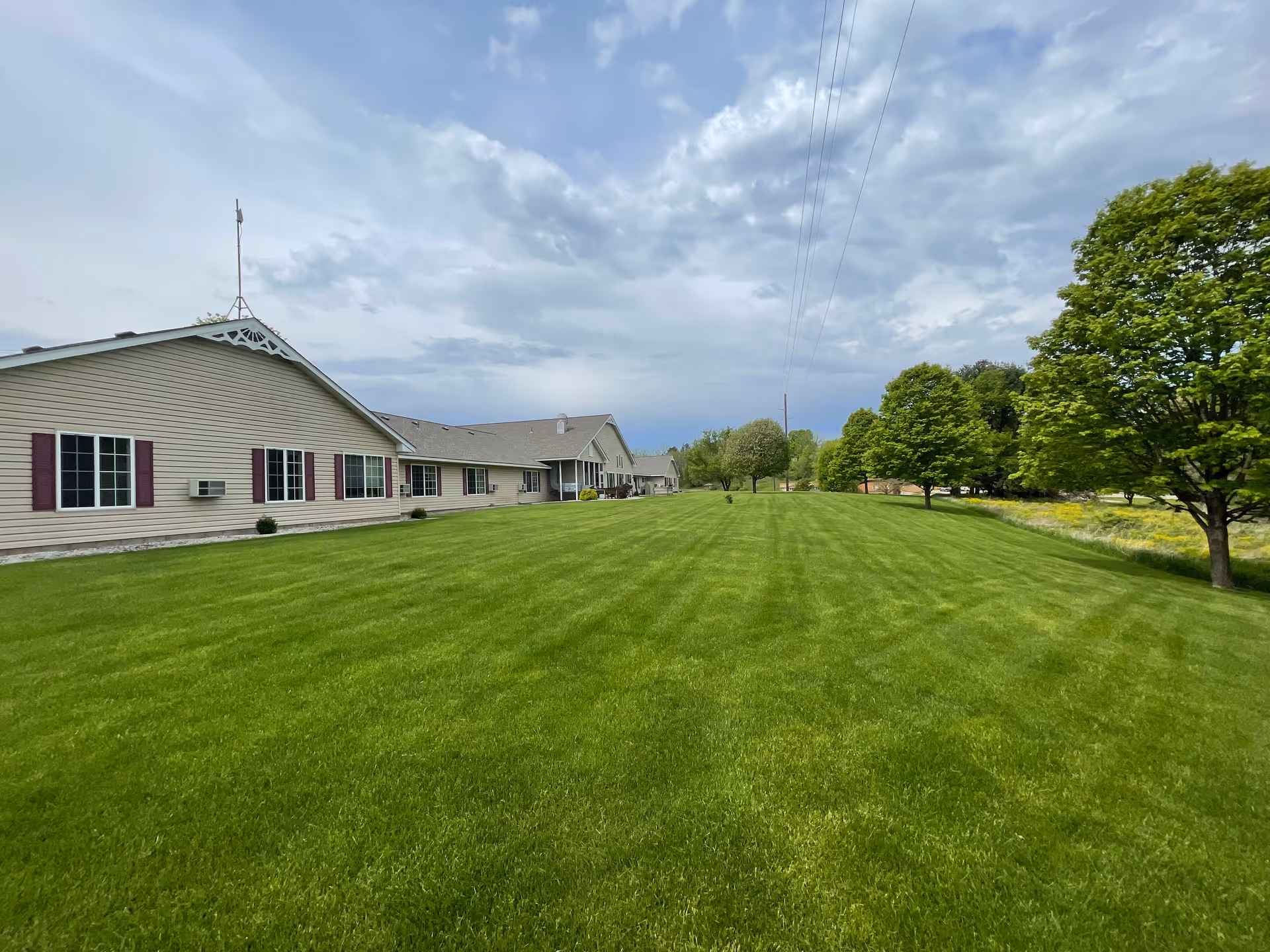 Wide view of a large green lawn with neatly mowed grass in front of a long, single-story beige building with multiple windows and maroon shutters. Several trees line the right side of the lawn under a cloudy sky.