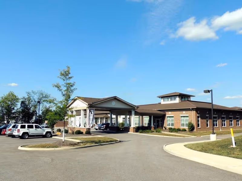 Front entrance of a single-story brick senior care facility with a porte-cochère, parking lot, and clear blue sky.