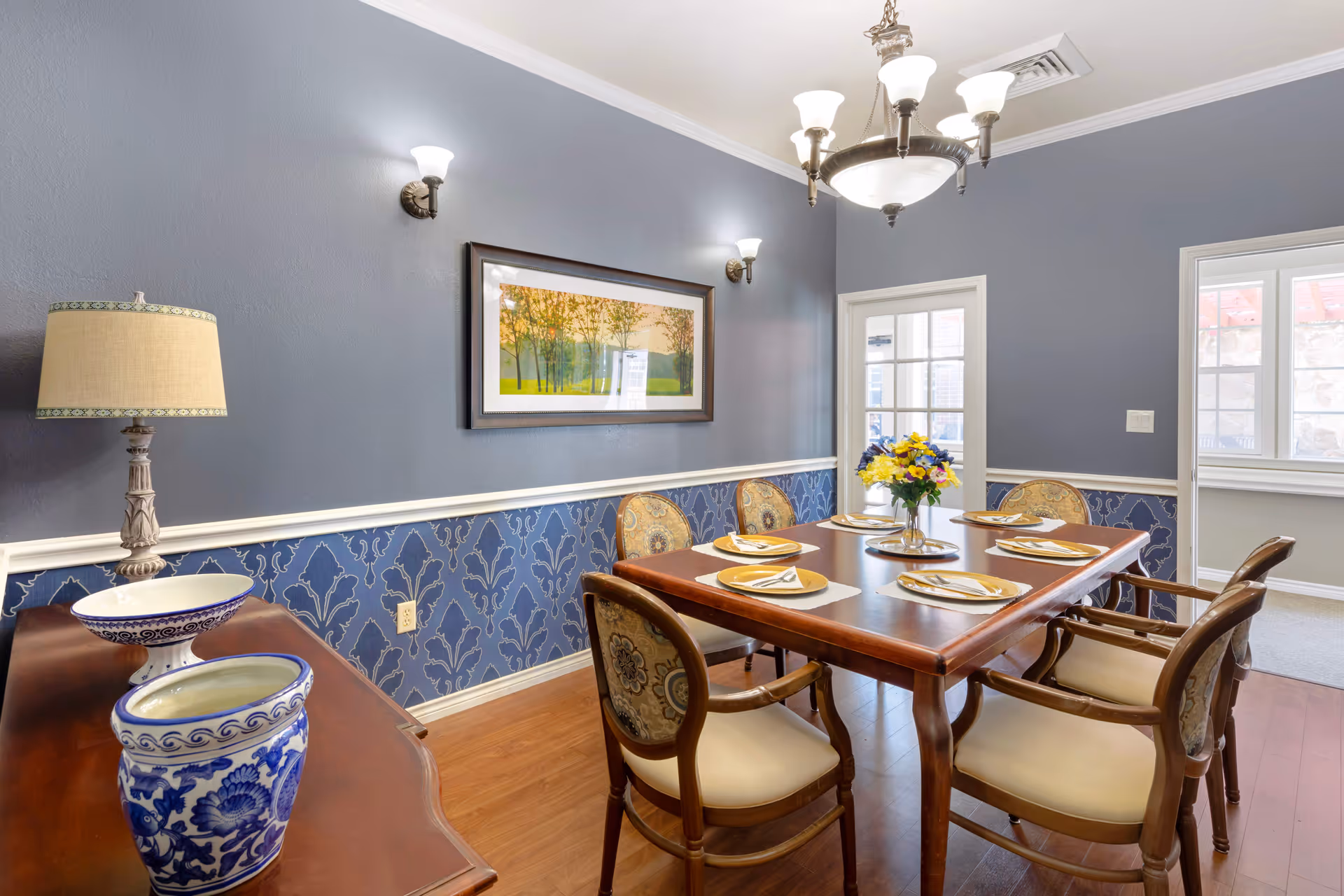 A dining room with a wooden table set for six people, each place setting having a plate, napkin, and utensils. The chairs have wooden frames with cushioned seats and backs featuring a floral pattern. The walls are painted blue with a decorative wallpaper border on the lower half. A framed landscape painting hangs on the wall above the table. There is a sideboard with a lamp and decorative blue and white ceramic pieces. The room is lit by wall sconces and a chandelier, and there are windows and a glass door letting in natural light.