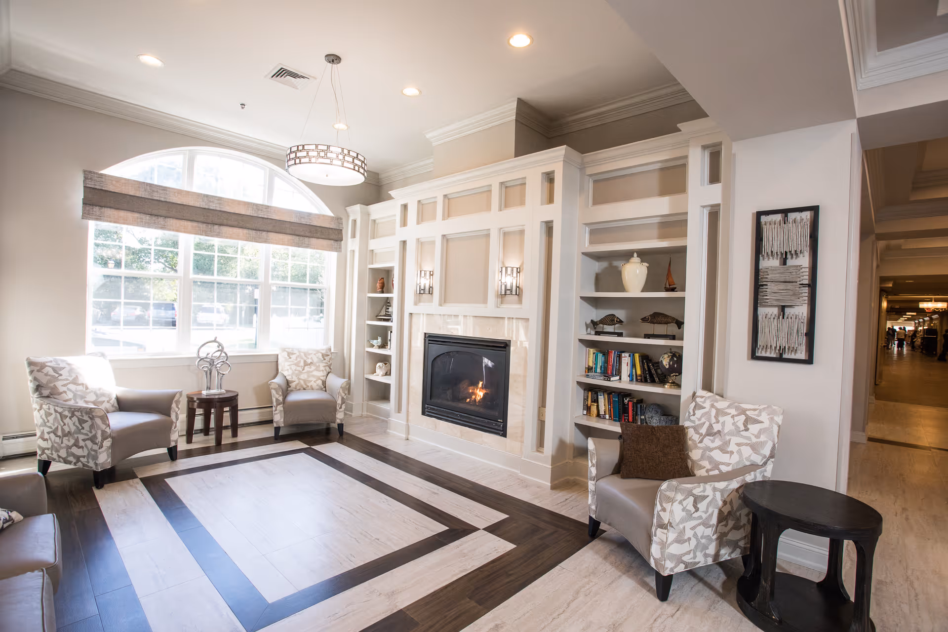 Bright communal living room with a fireplace, built-in shelves, patterned armchairs and a large arched window.