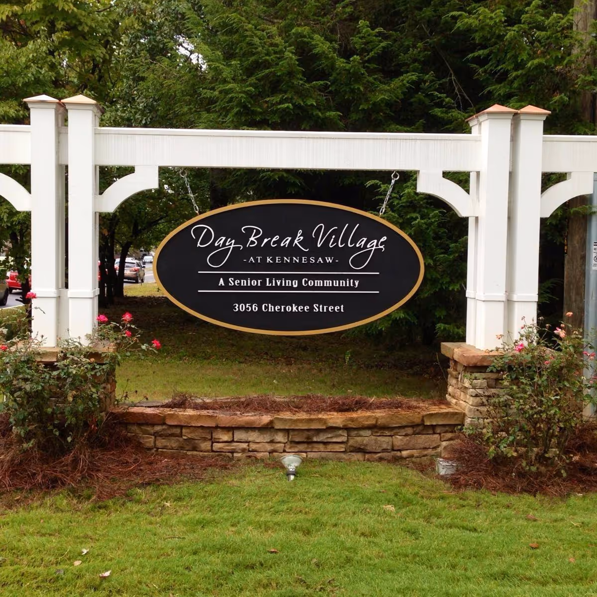Entrance sign for Day Break Village senior living community hanging from a white wooden frame amid landscaping.