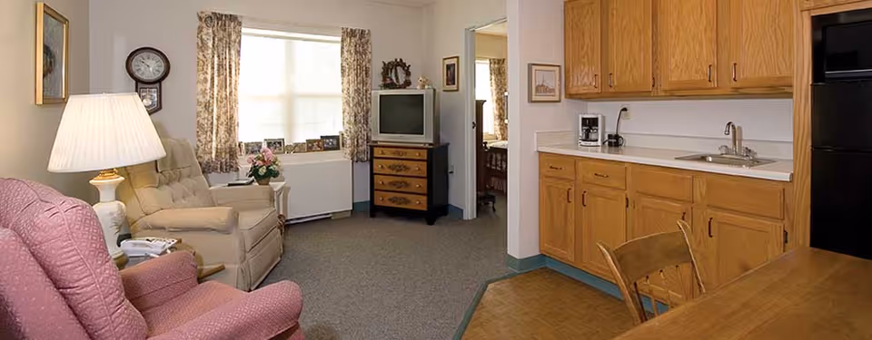 Sunlit senior living room with upholstered chairs, a TV on a dresser, and a small kitchenette with oak cabinets and sink.